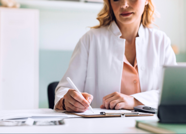 Doctor in a white coat writing on a clipboard at her desk. 